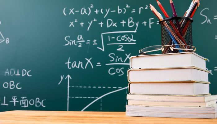 A stack of books with pencil holder and glasses against a chalkboard